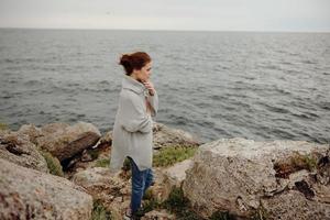 mooi vrouw in een grijs trui staat Aan een rotsachtig kust natuur vrouw ontspannende foto