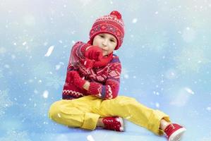 kind in warm kleren Aan een blauw winter achtergrond. een jongen in een rood gebreid hoed en trui. modieus jongen in de studio in wit sneeuwvlokken foto