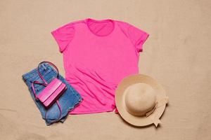 zand strand structuur achtergrond. mockup roze zomer t-shirt kleding kopiëren ruimte. blanco sjabloon vrouw overhemd top visie. zomertijd accessoires hoed, jeans. plat leggen detailopname t-shirt Aan kust. strand tijd foto