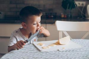 schattig jongen 4-5 jaren oud met mes snijdend een kaas Aan de snijdend bord Aan de tafel in de keuken. foto