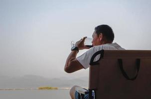 Mens zittend Aan een stoel en drinken rood wijn door de meer. foto