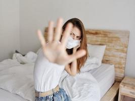 roodharig vrouw in een medisch masker is gedekt door haar handen van de camera terwijl zittend Aan de bed binnenshuis foto