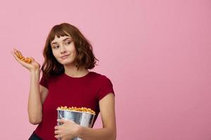 opgewonden verward schattig roodharige dame in rood t-shirt met popcorn klaar voor film avond poseren geïsoleerd Aan over- roze studio achtergrond. kopiëren ruimte spandoek. mode bioscoop concept. vermaak aanbod foto