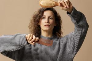 lief gelukkig gekruld mooi vrouw in grijs gewoontjes trui houden haarborstel kam in voorkant van camera poseren geïsoleerd Aan over- beige pastel achtergrond. problematisch onhandelbaar beschadigd haar- concept. kopiëren ruimte foto