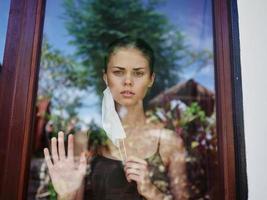 vrouw in de venster vervelend een medisch masker verdrietig kijken foto