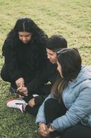 drie mensen van Spaans-Latino etniciteit, zittend Aan de grond in de park met slim apparaten foto