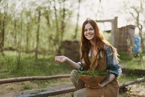 een jong vrouw feeds haar kippen Aan de boerderij met gras, vervelend een gemakkelijk plaid shirt, broek en schort, en glimlachen voor de camera, zorgzaam voor de dieren foto