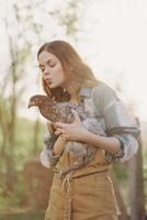 een gelukkig jong vrouw glimlacht Bij de camera en houdt een jong kip dat legt eieren voor haar boerderij in de zonlicht foto