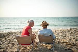 een paar van ouderen zittend in stoelen Bij de strand aan het kijken de zon en de zee Aan hun zomer vakantie en ze glimlach en genieten hun vakantie. foto