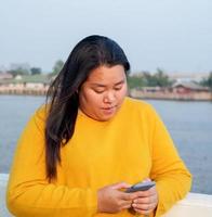 portret dik Aziatisch vrouw lang zwart haar- vervelend geel overhemd zijn gebruik makend van mobiel telefoon of smartphone werk met echt en echt gezicht en uitdrukking binnen park in avond uren zonsondergang dag foto