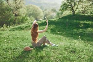 vrouw blogger zit Aan de groen gras in een park en duurt afbeeldingen van haarzelf Aan haar telefoon tegen de backdrop van een zomer landschap. jong mensen levensstijl en bezorgdheid voor de milieu foto