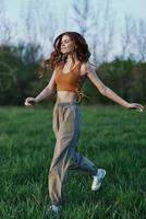 een jong vrouw in een top en groen broek en atletisch sportschoenen is jogging Aan de zomer groen gras met haar haar- los foto