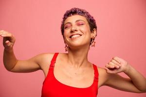 mode portret van een vrouw met een kort kapsel van Purper kleur en een glimlach met tanden in een rood top Aan een roze achtergrond dansen gelukkig foto