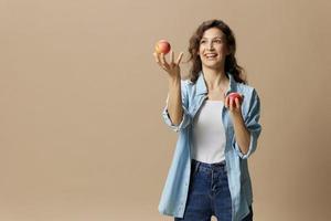 gelukkig glimlachen gefocust gekruld mooi vrouw in jeans gewoontjes overhemd jongleren met appel poseren geïsoleerd Aan over- beige pastel achtergrond. gezond voedsel. natuurlijk milieuvriendelijk producten concept. kopiëren ruimte foto