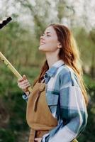 vrouw prachtig glimlachen boer in werk kleren en schort werken buitenshuis in natuur en Holding een hark naar verzamelen gras foto