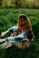 een jong, mooi vrouw lounges Aan de groen gras in de park vervelend sneaker broek en een plaid overhemd en looks uit in de instelling zon foto