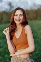 grappig jong roodharige vrouw in een oranje top glimlachen tegen de groen gras van natuur in de zonsondergang licht foto