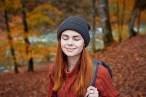 gelukkig vrouw met een rugzak reist in de park in herfst en gebaren met de handen van de rivier- in de achtergrond foto