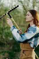 vrouw prachtig glimlachen boer in werk kleren en schort werken buitenshuis in natuur en Holding een hark naar verzamelen gras foto