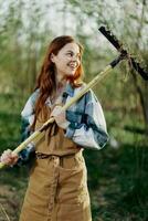 vrouw prachtig glimlachen boer in werk kleren en schort werken buitenshuis in natuur en Holding een hark naar verzamelen gras foto