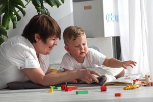 grootmoeder is aan het liegen Aan de verdieping met haar kleinzoon en aan het kijken een boek. de oppas is verloofd in ontwikkelen taken met een kleuter jongen. vroeg ontwikkeling. foto