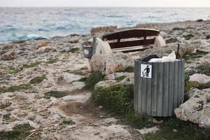 volle vuilnisbak of vuilnisbak met plastic fles, bierblikjes en organisch afval dat zichtbaar vervuiling vertoont in kustgebieden bij de zee. houten bankje met emmer met afval op rotsachtige kust.