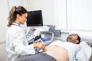 voorzichtig vrouw dokter in wit jas zittend in voorkant van een echografie inrichting en dirigeren buik diagnostiek met transducer foto