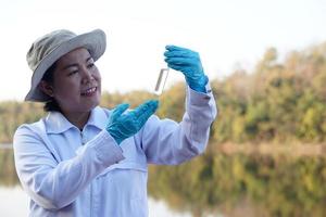 Aziatisch vrouw milieu onderzoeker houdt buis van monster water naar inspecteren van de meer. concept, ontdekken, analyse water kwaliteit van natuurlijk bron. ecologie veld- Onderzoek. foto