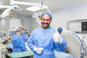 jong mannetje chirurg staand in in werking kamer tonen duimen omhoog, klaar naar werk Aan een geduldig. mannetje medisch arbeider chirurgisch uniform in operatie theater. foto