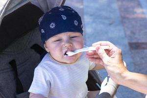 een meisje moeder feeds haar weinig jongen zoon van een lepel Aan de straat. foto