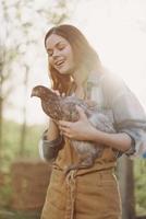 een gelukkig jong vrouw glimlacht Bij de camera en houdt een jong kip dat legt eieren voor haar boerderij in de zonlicht foto
