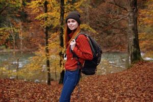 vrouw in herfst park met gedaald bladeren en rugzak Aan haar terug rivier- in de achtergrond foto