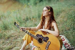 hippie vrouw eco vriendelijk spelen gitaar glimlacht en het zingen liedjes in natuur zittend Aan een plaid door de meer in de avond in de zonsondergang zonlicht. een levensstijl in harmonie met de lichaam en natuur foto