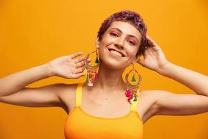 portret van een jong vrouw met een kort kapsel en gekleurde haar- glimlachen en tonen haar tong Bij de camera Aan een oranje achtergrond met oorbellen accessoires in de studio foto