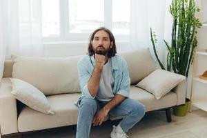 een freelance Mens in een wit t-shirt, blauw jeans, en overhemd zit Aan de bankstel met zijn telefoon Bij huis Aan zijn dag uit en Toneelstukken spellen foto