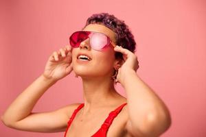 mode portret van een vrouw met een kort kapsel in gekleurde zonnebril met ongebruikelijk accessoires met oorbellen glimlachen Aan een roze helder achtergrond met een geschiktheid lichaam dansen foto