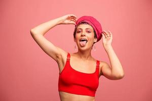 jong atletisch vrouw met een kort kapsel en Purper haar- in een rood top en een roze hoed met een atletisch figuur glimlacht en grimassen op zoek Bij de camera Aan een roze achtergrond foto