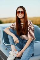 een modieus vrouw glimlacht zoet in elegant zonnebril, een gestreept t-shirt en jeans, zit Aan de romp van een auto en looks Bij de mooi natuur van herfst. reizen levensstijl foto