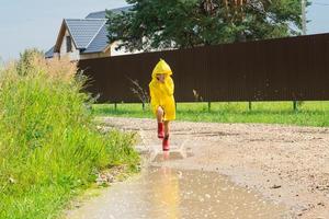 een meisje in rood rubber laarzen en een geel regenjas loopt door plassen na een regen in de dorp. zomer tijd, vrijheid, kinderjaren foto