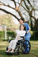 ouderen Aziatisch senior vrouw Aan rolstoel met Aziatisch voorzichtig verzorger en aanmoedigen geduldig, wandelen in tuin. met zorg van een verzorger en senior Gezondheid verzekering. foto