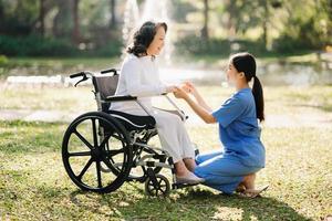 ouderen Aziatisch senior vrouw Aan rolstoel met Aziatisch voorzichtig verzorger. verpleging huis ziekenhuis tuin concept. in zon licht foto