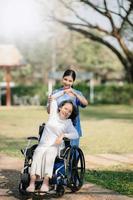 Aziatisch voorzichtig verzorger of verpleegster en de gelukkig geduldig in een rolstoel zijn wandelen in de tuin naar helpen en aanmoedigen en rust uit uw geest met groen natuur. helpen ondersteuning jezelf naar leren naar wandelen. foto