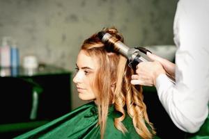 kapper gebruik makend van curling tang krullen lang bruin haar- Aan de jong Kaukasisch meisje in een schoonheid salon. foto