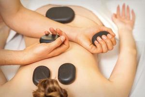 heet steen massage Aan de vrouw terug met handen van masseur Holding zwart massage stenen in spa salon. foto