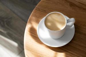 een wit porselein kop met koffie Aan een schotel staat Aan een ronde tafel foto