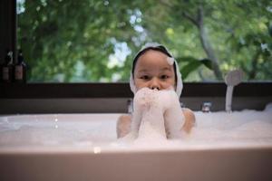 portret van een Aziatisch weinig schattig meisje met geluk en pret spelen met lichaam zeep schuim, shampoo plons, en water bubbels in een wit bad Bij een hotel badkamer met groen boom venster achtergrond. foto