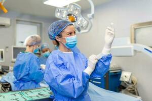 portret van mooi vrouw dokter chirurg zetten Aan medisch handschoenen staand in operatie kamer. chirurg Bij modern in werking kamer foto