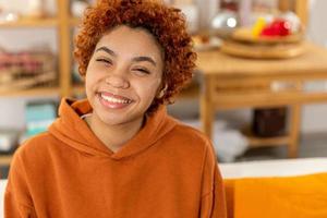 mooi Afrikaanse Amerikaans meisje met afro kapsel glimlachen zittend Aan sofa Bij huis binnen. jong Afrikaanse vrouw met gekruld haar- lachend. vrijheid geluk zorgeloos gelukkig mensen concept. foto