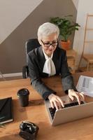 zelfverzekerd elegant Europese midden- oud senior vrouw gebruik makend van laptop Bij werkplek. elegant ouder volwassen Jaren 60 grijs haren dame zakenvrouw zittend Bij kantoor tafel. baas leider leraar professioneel arbeider. foto