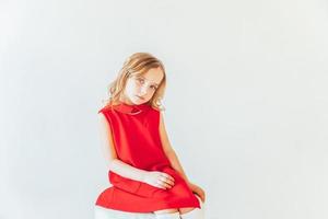 zoet weinig meisje in rood jurk zittend Aan stoel tegen wit muur Bij huis, ontspannende in wit helder leven kamer binnenshuis. kinderjaren schoolkinderen jeugd kom tot rust concept. foto